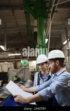 Due tecnici nelle offerte di lavoro e di usura in hardhats guardando blueprint e discuterne insieme in fabbrica Foto Stock