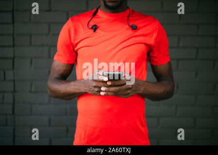 Giovane uomo sportive in piedi di fronte a un muro di mattoni, utilizzando il suo smartphone Foto Stock