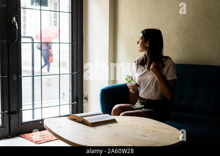 Giovane donna con un telefono cellulare seduta su un divano guardando fuori della finestra Foto Stock