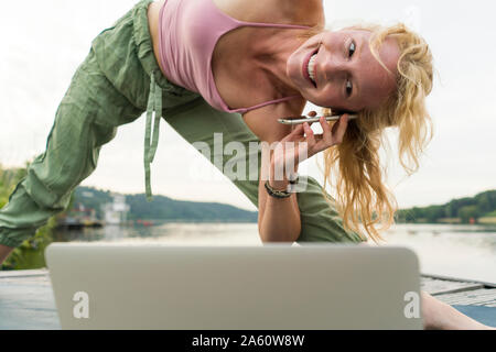 Giovane donna stretching e utilizzando computer portatile e un telefono cellulare su un pontile a lago Foto Stock