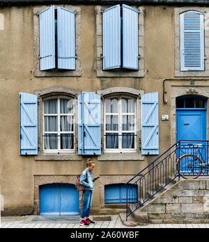 Saint-Vaast-la-Hougue, Manche / Francia - 16 Agosto, 2019: ragazza vestita di azzurro che interagisce con il suo telefono cellulare nella parte anteriore di una casa con blue s Foto Stock