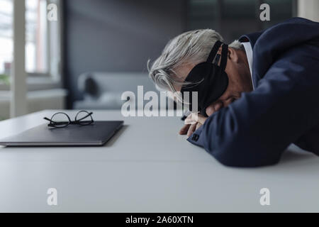 Imprenditore Senior che indossa la maschera per gli occhi sdraiato sulla scrivania Foto Stock