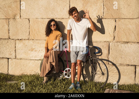 Coppia giovane con la bicicletta, appoggiata sul muro di pietra, facendo segni a mano Foto Stock