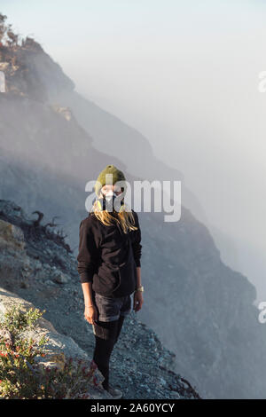 Giovane donna con maschera respiratore permanente al bordo del vulcano Ijen, Java, Indonesia Foto Stock