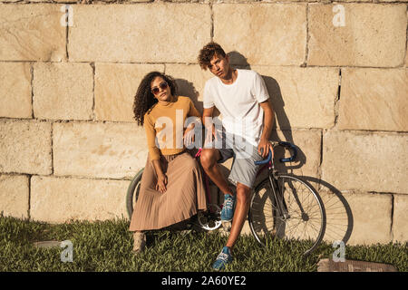 Coppia giovane con la bicicletta, appoggiata sul muro di pietra, alla moda Foto Stock