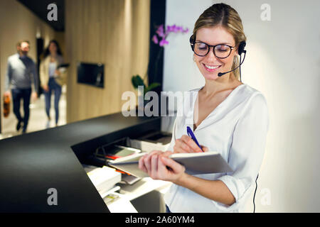 Un personale al ricevimento sorridente parlando con cuffie mentre prendendo appunti Foto Stock
