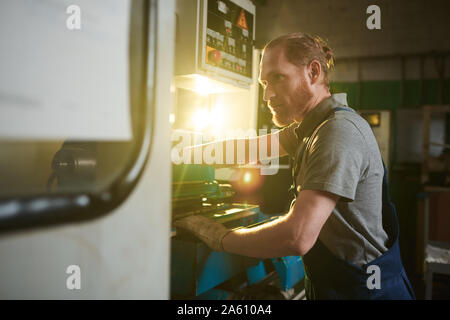 Lavoratore manuale in tute di concentrarsi sul suo lavoro da lui in piedi e lavorando al tornio in pianta Foto Stock