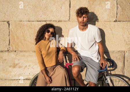 Coppia giovane con la bicicletta, appoggiata sul muro di pietra, alla moda Foto Stock