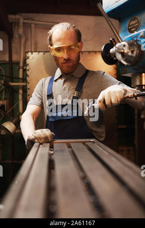 Barbuto giovane lavoratore manuale in occhiali protettivi lavora con metallo su attrezzature speciali in fabbrica Foto Stock