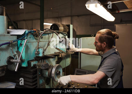 Giovanissimo meccanico di usura di lavoro in piedi vicino al tornio e a lavorare in fabbrica Foto Stock