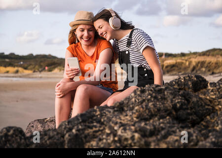 Due amiche seduto sulla spiaggia rocciosa, utilizza lo smartphone, ascolto miusic Foto Stock