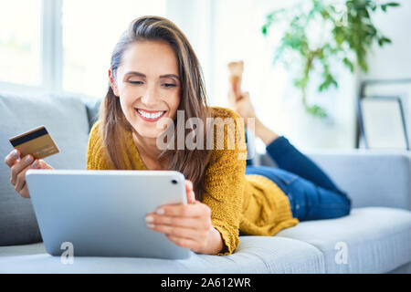Allegro giovane donna utilizzando la carta di credito e tablet per shop online da casa Foto Stock