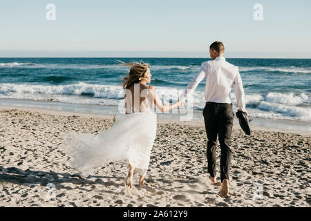 Vista posteriore della sposa e lo sposo in esecuzione sulla spiaggia Foto Stock