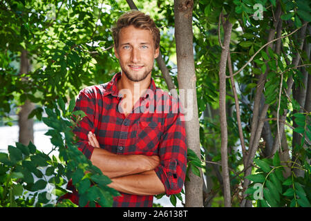 Ritratto di fiducioso giovane uomo che indossa camicia a scacchi appoggiato a un albero Foto Stock