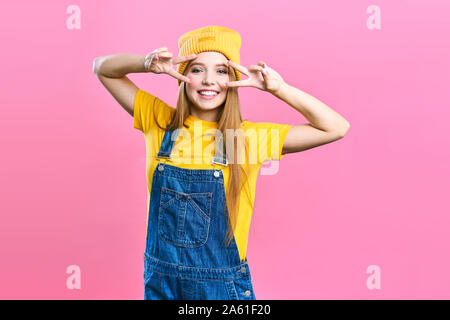 Portret una bella ragazza in tuta in jeans e un cappello giallo su sfondo rosa. Fashionista signora sorridente dello studente . Luminose alla moda fashion studio immagine Foto Stock