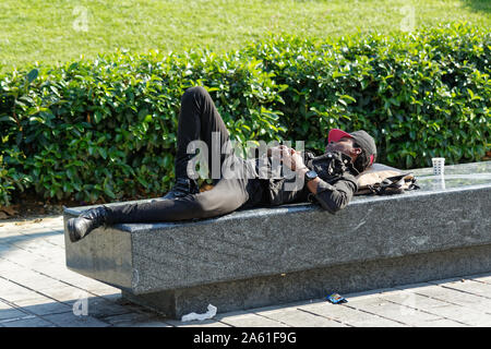 Beyoglu, Turchia - 20 Ottobre 2019: giovane africano con cappello da baseball e abiti neri di dormire su una panca in calcestruzzo a parco pubblico di Istanbul Foto Stock