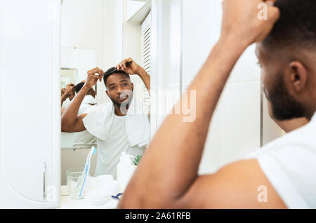 Giovane uomo nero esaminando i suoi capelli, guardando lo specchio del bagno Foto Stock