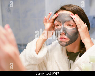 Cosmetici maschera nera. La donna porta la bellezza per il suo volto si sbarazza di puntini neri, utilizzando una maschera nera. Una femmina in bagno guarda nello specchio Foto Stock