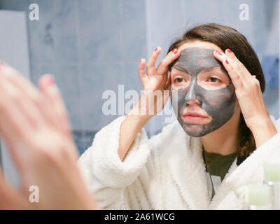 La ragazza in maschera facciale guarda nello specchio infastidetto. Cosmetici maschera nera. La donna porta la bellezza per il suo volto si sbarazza di puntini neri, utilizzando un nero Foto Stock