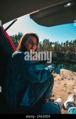 Matura nel baule auto con bellissima vista lago. prospettiva in prima persona. giovane donna sorridente Foto Stock