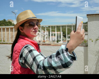 Giovani attraenti brunette in un cappello assume un selfie dal suo bianco telefono cellulare Foto Stock