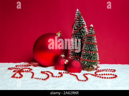 Decorazione di natale con tema di piccole dimensioni e decorata di abeti rossi e baubles di perline sulla neve contro lo sfondo di colore rosso. Merry Christmas greeting card. Foto Stock