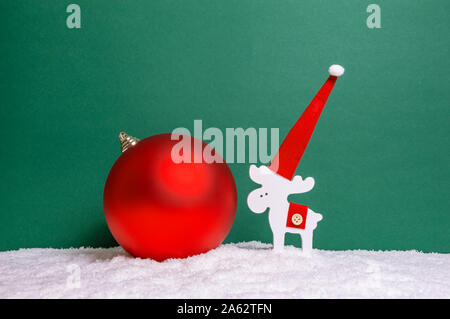 Minimalista per le vacanze di Natale con il concetto di legno bianco in renna Santa's hat e big red ninnolo sulla neve contro lo sfondo di colore verde. Buon Natale gr Foto Stock