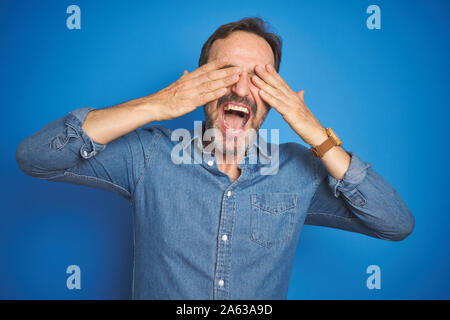 Bella la mezza età senior uomo con i capelli grigi isolati su sfondo blu che copre gli occhi con le mani sorridendo allegro e divertente. Concetto di ciechi. Foto Stock