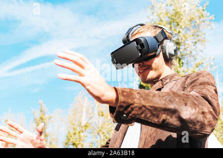 Giovane maschio con i capelli lunghi che indossa occhiali per realtà virtuale e grandi cuffie per esterni. Smartphone con occhiali VR auricolare. Foto Stock
