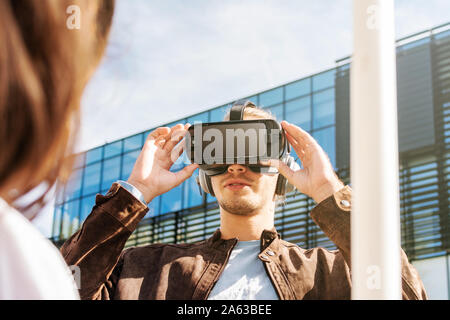 Giovane maschio con i capelli lunghi che indossa occhiali per realtà virtuale e grandi cuffie per esterni. Smartphone con occhiali VR auricolare. Tenendo i bicchieri con la rai Foto Stock
