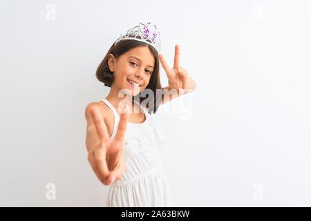 Bella ragazza bambino indossando princess crown in piedi isolato su sfondo bianco sorridente guardando alla telecamera che mostra le dita facendo segno di vittoria. Foto Stock