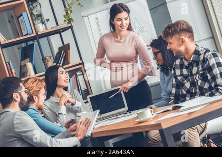 Startupers lavorando insieme a Office seduta guardando la donna incinta in piedi rivolta verso lo schermo del notebook gioiosa Foto Stock