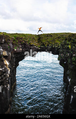 Tourist salta sopra un ponte naturale di roccia in Arnarstapi, Islanda Foto Stock