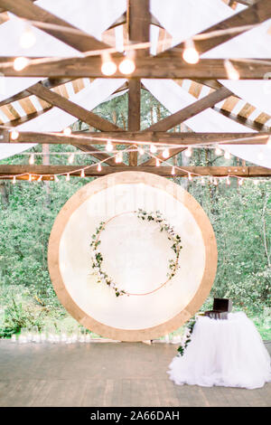 Nozze arco tondo in stile rustico decorato con ghirlande di fiori e vegetazione, retro lampadine sul soffitto. Bellissimo matrimonio istituito. Area Foto Stock