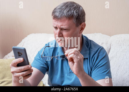 Un uomo con gli occhiali guarda a uno smartphone mentre è seduto in un comodo divano. Di mezza età imprenditore controlla la posta in uno smartphone Foto Stock