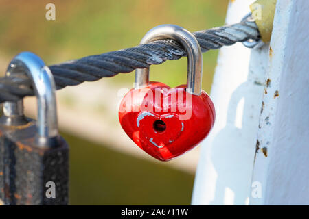 Il dispositivo di bloccaggio rosso appendere su un cavo su un ponte. Simbolo di amore e fedeltà eterna metal heart-a forma di lucchetto sul ponte di close-up. Tradizioni di matrimonio. Foto Stock