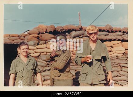 Tre giovani soldati americani, un gruppo multirazziale con due uomini caucasici e un uomo afro-americano, sorridono mentre si posano davanti ai sacchi di sabbia in una fortificazione in Vietnam durante la guerra del Vietnam, 1975. () Foto Stock