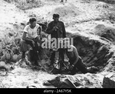 Colpo di alto angolo di due soldati americani, in piedi fuori in un dugout sabbioso, guardando un collega inginocchiato a terra per guardare attraverso le viste del treppiede cannone montato o telescopio, Vietnam, 1965. () Foto Stock