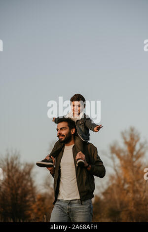Padre Felice con il figlio sorridente sulle sue spalle a camminare nel parco d'autunno. Foto Stock