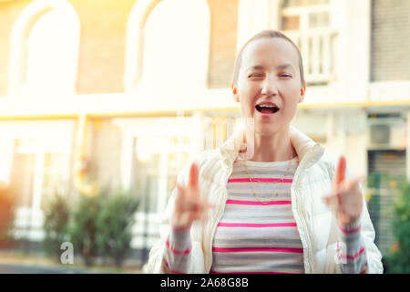 Felice caucasian giovane adulto hipster ragazza ritratto winking e mostrando thums fino firmare all'aperto a caldo la sera al tramonto in autunno. Funky bella donna Foto Stock