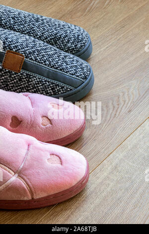 Due coppie di casa pantofole, mens grigio e rosa womens su un marrone con pavimento in legno. Accoglienti e caldi e confortevoli scarpe da interno. Vista laterale. Foto Stock