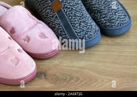 Due coppie di casa pantofole, Womens colore rosa e grigio mens su un marrone con pavimento in legno. Accoglienti e caldi e confortevoli scarpe da interno. Vista laterale. Foto Stock