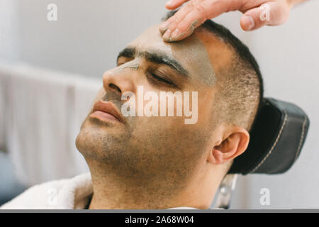 La pelle maschile cura nel salone di bellezza. Applicazione di argilla Maschera di pulizia su una il volto dell'uomo Foto Stock