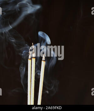 Tre bastoncini di incenso bruciare con emissione di fumo bianco su marrone scuro sfondo, culto culto in credenze asiatiche e fede Foto Stock