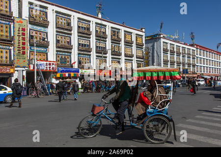 Un pedicab porta un passeggero di fronte appartamenti moderni con disegni tradizionali a Lhasa, in Tibet. Foto Stock