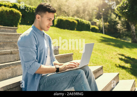 Per il tempo libero all'aria aperta. Elegante ragazzo indossa guarda seduti sulle scale in Parco lavorando su laptop concentrata vista laterale Foto Stock