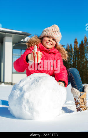 Vacanza invernale. Ragazza in piedi all'aperto vicino casa seduta di rotolamento per le palle di neve pupazzo di neve sorridente allegro close-up Foto Stock