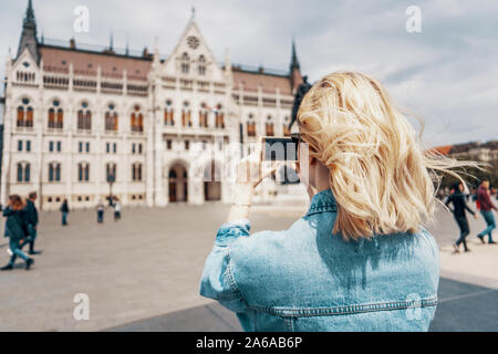 Giovane turista femminile prende le immagini del parlamento ungherese con il suo smartphone sulla bella giornata di sole. Inquadratura da dietro. Unione le destinazioni di viaggio Foto Stock