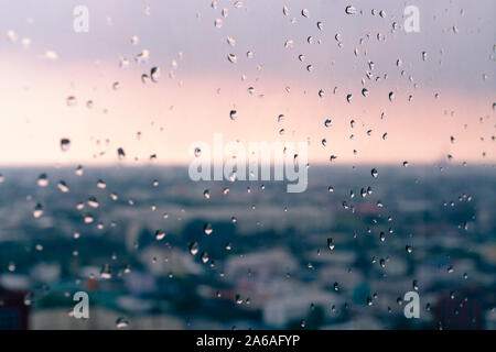 Gocce di pioggia sulla finestra appartamento con vivaci città tramonto sfondo sfocato - le goccioline di acqua sul grattacielo piano di vetro con urban golden ora sky - Meteo, umore e il concetto di stagione Foto Stock