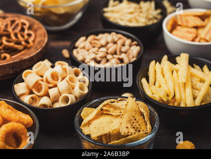 Tutti i classici snack di patate con arachidi, popcorn e anelli di cipolla e salati pretzel nella ciotola piastre su legno. Ghirigori con bastoni e patatine e cr Foto Stock